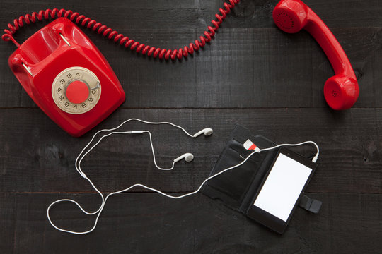 The image shows a hooked off red old retro phone  with a smartphone with white earphones on wooden background