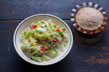 Vegetable salad made of fresh cabbage, radish, cucumber, bell pepper and sour cream in a white plate and sea salt with seasoning in a glass shaker on the old black table. Top view closeup