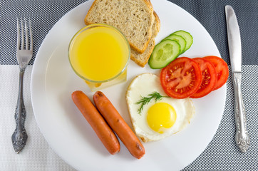 breakfast on a white plate on a black and white table Fried egg in a heart, fried sausage, fresh vegetables, juice, sliced bread, knife and fork on either side, napkin