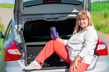 Mature woman drinking a drink sitting on the trunk of a car