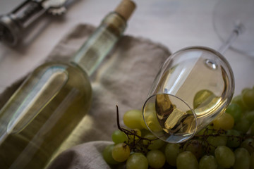 wineglass with white wine with bottle and grapes on white wood textured table covered with canvas...