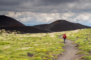 Schilderijen op glas Lonely unrecognizable Hiker dressed in in red walks along a path on Mount Etna in Sicily, heading to a black volcano crater. © AMzPhoto