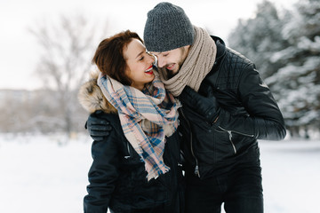 Cute young hipster couple having fun in winter park on a bright day hugging each other and smiling
