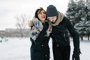Cute young hipster couple having fun in winter park on a bright day hugging each other and smiling
