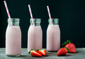 Smoothie milkshake made from fresh strawberry blended with kefir yogurt. Served in bottle style glasses and with fresh strawberries. Served on a stone slate table with colorful stripy straws.