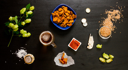 Beer set. Glass of beer, crackers, ketchup, salt. The ingredients for brewing: malt and hops. On a wooden black background.