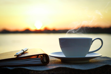 cup of hot coffee with a pen and note book in sunrise background over the river