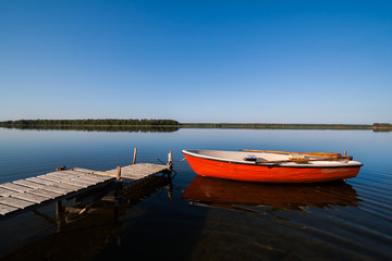 A beautiful idyllic view of a glassy lake in northern Sweden. The sky is perfectly blue and you see a rowing boat with fishing gear docked by a small jetty.
