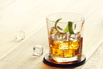 Glass of whiskey with ice and lime peel on wooden background, warm color tone