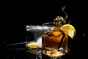 Falling piece of ice into quality glass of whiskey makes very many splashes and other glass lies on side with lemon slice, tubules and ice on black background.