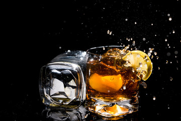 Two high quality glasses of whiskey, one lies on side and other with lemon slices and ice and many splashes on black background.