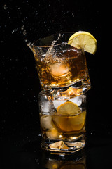 Falling piece of ice in one high quality glass of whiskey with splashes standing on another glass full of ice and lemon slice on black background.