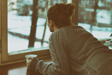 Sad woman by the window, looking out, with a cup of coffee in a hand