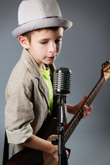 Little boy playing guitar and singing with microphone on grey background