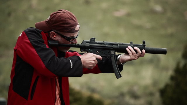 Man in baseball cap and red jacket shooting an MP5
