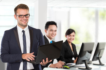 Young caucasian businessman, with his team behind holding laptop