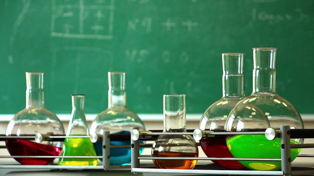 Six glass flasks filled with solutions of different colors synchronously rotate on the laboratory shakers on the green chalkboard background. Focus on right (front) platform. No camera movement. 