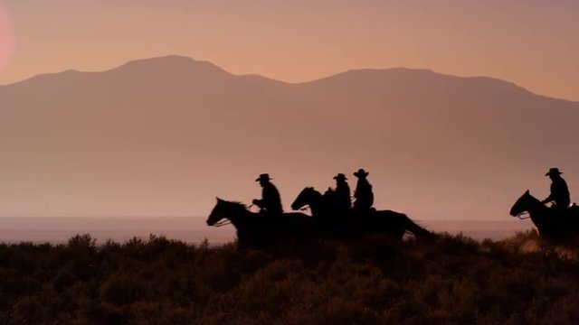 Cowboys galloping riding horses silhouetted against orange mountains in slow motion.