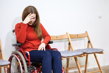 Disabled crying woman in wheelchair covers her face with hand while sitting between chairs