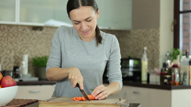 Young smiling woman cutting carrots on kitchen counter board