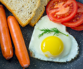 Breakfast fried egg in heart-shaped, grilled sausages, tomatoes, bresd, top view, dark background