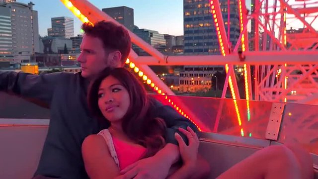 An attractive couple cuddled together on a ferris wheel at night, city buildings behind them