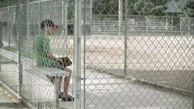 Slow motion push of boy sitting in dugout behind chain link fence.