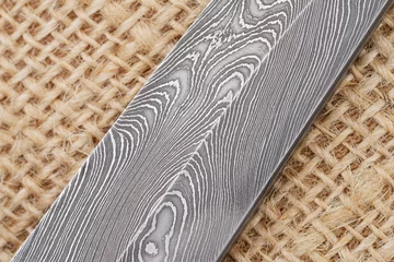 Tuinposter Fragment of the traditional handmade Finnish knife blade with the abstract wave pattern of damascus steel over an old sack background. © Dmitry Chulov