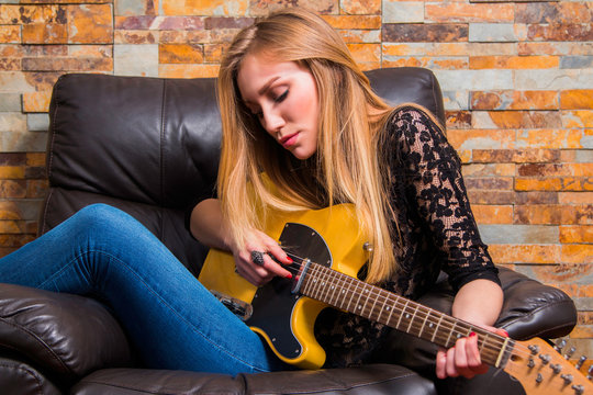 Attractive young girl in blue jeans and black lace shirt sitting in chair and playing guitar on brick background