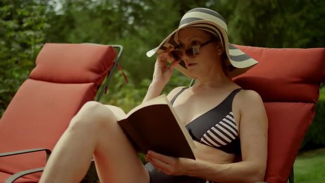 An older woman reading by the pool sees something and gets up, in slow motion