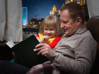 Отец читает дочке книжку. Папа с маленькой дочкой в кресле у окна. Дедушка и внучка. За окном ночной город, Москва. Вечер. Чтение на ночь, перед сном.