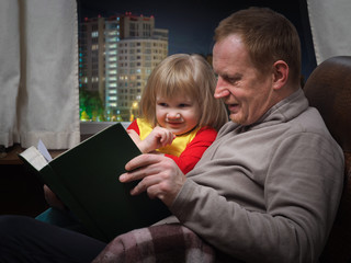 Отец читает дочке книжку. Папа с маленькой дочкой в кресле у окна. Дедушка и внучка. За окном ночной город, Москва. Вечер. Чтение на ночь, перед сном.