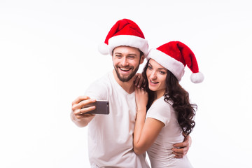 Obraz premium Christmas young beautiful couple in Santa hats in love taking romantic self portrait selfie photo together with mobile phone smiling happy wearing on white background