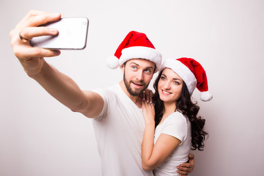 christmas, holidays, technology and people concept - happy couple in santa hats taking selfie picture with smartphone on white background
