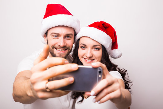 christmas, holidays, technology and people concept - happy couple in santa hats taking selfie picture with smartphone on white background