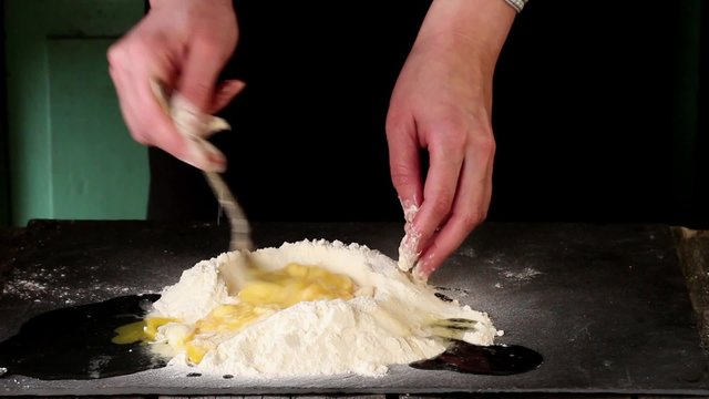 Female hands knead eggs in flour for making dough over black table