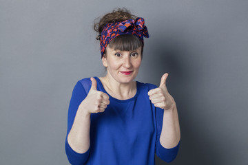 optimism concept - shy retro 30s woman smiling with two thumbs up for embarrassed success, studio grey background