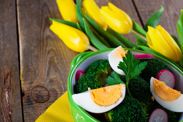 Salad with broccoli, eggs and radishes in a small bowl on wooden background and tulip bouquet, with copy space.