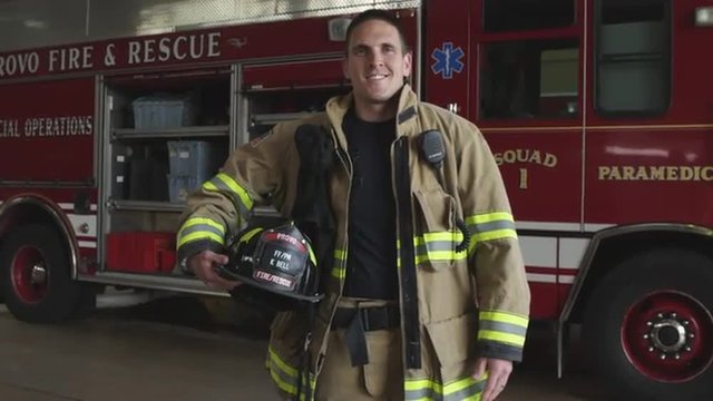 Fireman holding helmet in uniform, heroric and smiling next to fire truck in station.