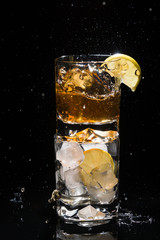 One high quality glass of whiskey with splashes standing on another glass full of ice and lemon slice on black background.