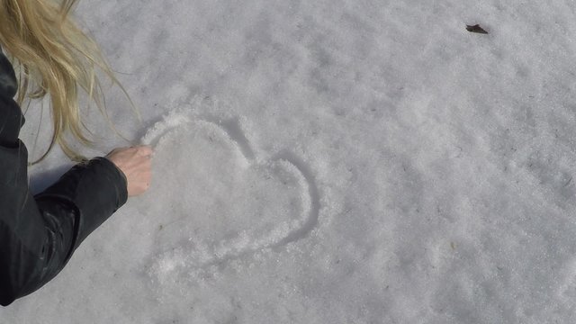 Blonde woman hand drawn heart symbol on snowy ground at sunny day.