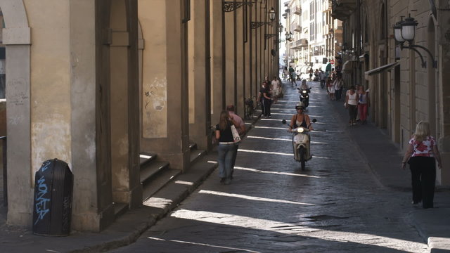People and scooters passing by an old archway in Italy.