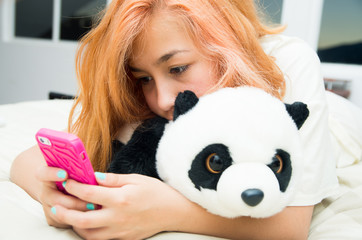 Pretty young woman horizontally lying cozy in bed using pink mobile phone and stuffed panda animal