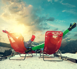 Two happy skiers sitting in chaise-longues on the top of snow mo