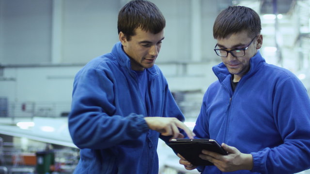 Two aircraft maintenance mechanics have a conversation while using a tablet in a plane hangar. Shot on RED Cinema Camera.