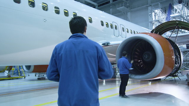Aircraft maintenance mechanic in blue uniform is walking to greet his colleague who is checking the plane turbine blades in a hangar. Shot on RED Cinema Camera.