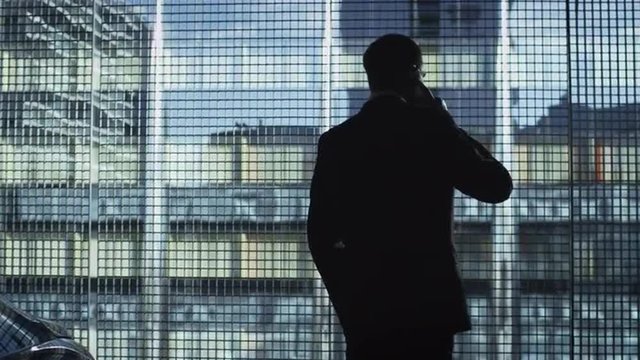 Businessman in a suit is talking on a phone next to an executive car and a big window in a garage. Shot on RED Cinema Camera.
