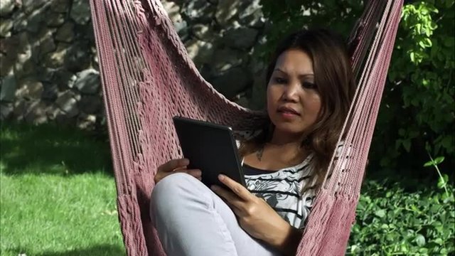 Handheld shot of an Asian woman reading something in her tablet while sitting on a hammock