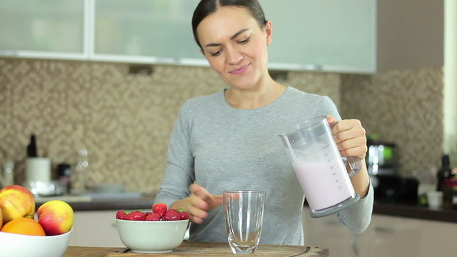 Cheerful brunette woman with blender and strawberries pouring milk shake to glass at home, drinking and smiling in front of camera