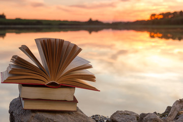 Stack of books and Open hardback book on blurred nature landscape backdrop against sunset sky with...